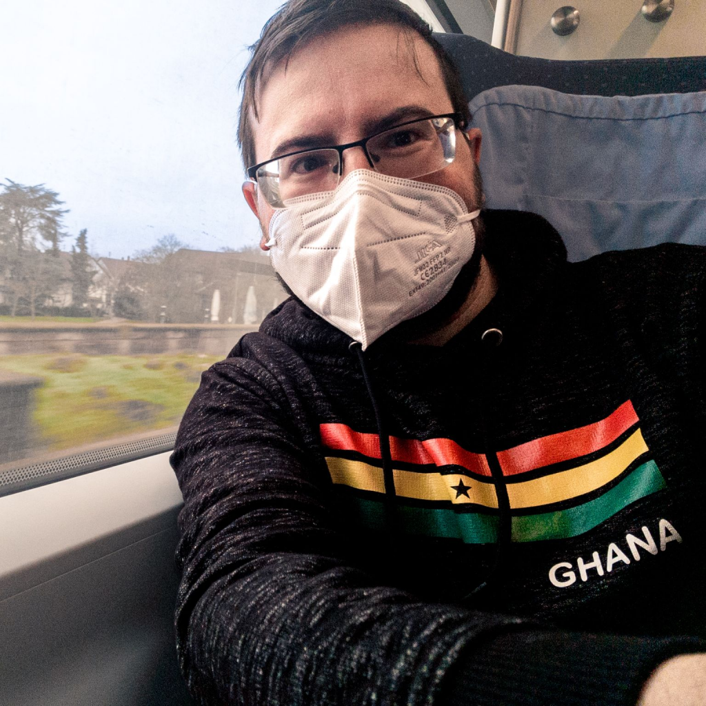 The image shows a person sitting on a train or bus, wearing a face mask and glasses. The individual is dressed in a black hoodie with colorful horizontal stripes in red, yellow, and green, and the word "GHANA" written on the sleeve. The background outside the window appears to be a scenic view with trees and a body of water, suggesting the person is traveling through a picturesque area. The person is looking directly at the camera, giving a thumbs-up gesture with their right hand.
