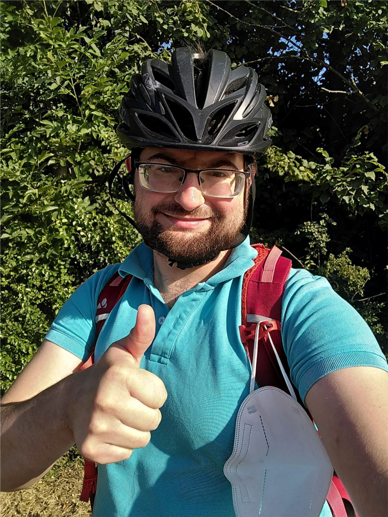 A person wearing glasses, a black bicycle helmet, and a light blue polo shirt is standing outdoors with greenery in the background. They have a red backpack on and a white face mask hanging from one of the backpack straps. The person is smiling and giving a thumbs-up gesture.
