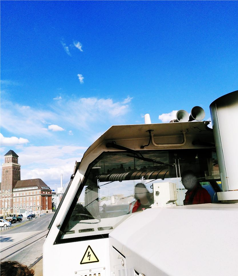 The image shows the front section of a shunting locomotive with a clear blue sky and a few clouds in the background. The vehicle's windshield is visible, and you can see the interior controls and two blurred individuals inside, likely the driver and another person. There is a yellow triangular warning symbol with a lightning bolt, indicating high voltage or electrical hazard, on the exterior of the vehicle. In the background, there are buildings, including a distinctive tower structure, suggesting an urban environment. The scene appears to be during daytime.