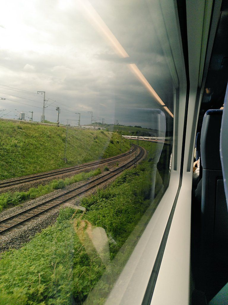 The image shows a view from inside a train, looking out through the window. The scene outside features railway tracks curving gently to the right, surrounded by green vegetation and grassy embankments. Overhead electric lines and poles are visible, indicating an electrified railway system. The sky is overcast with gray clouds, suggesting a cloudy day. The reflection in the window shows part of the train's interior and a person holding a phone.