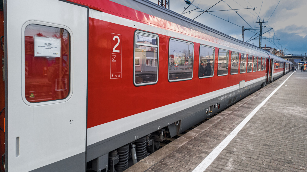 The image shows a red and white passenger train at a railway station. The train has a modern design with large windows, allowing a view of the interior where passengers are seated. The train car visible in the image is marked with the number "2" and the seats "12-78." There is a sign on the window indicating the train route, which includes stops Wendingen, Merklingen, Ulm. The platform is paved with bricks and has a white tactile strip along its edge for safety. Overhead electric lines and poles are visible, indicating that the train is powered by electricity. The sky is partly cloudy, suggesting a typical day.