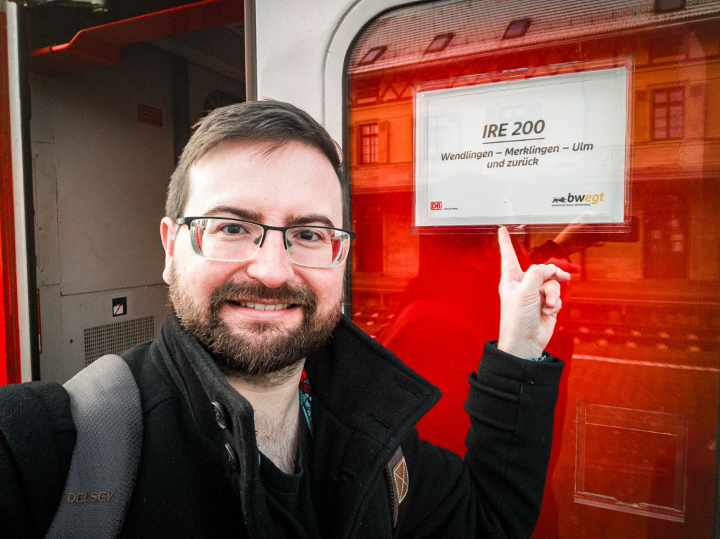 The image shows me standing at the door of a train. The individual is wearing glasses, has a beard, and is smiling at the camera. They are dressed in a dark jacket and have a backpack on their shoulder. The person is pointing towards a sign on the train window that reads "IRE 200" with destinations listed as "Wendlingen - Merklingen - Ulm und zurück," indicating the route of the train. The train interior is visible, with red and grey tones, and the person appears to be in a cheerful mood.